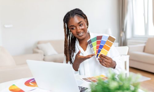 Cheerful african american lady designer having video conference with clients, sitting at desk in front of computer, holding color palettes, gesturing and smiling, copy space