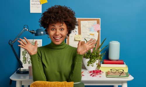 Intelligent female worker spreads palms, works in coworking space, has glad expression, dressed in green turtleneck, poses against blue background at desktop, has break after writing composition